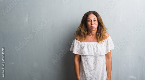 Middle age hispanic woman standing over grey grunge wall puffing cheeks with funny face. Mouth inflated with air, crazy expression.