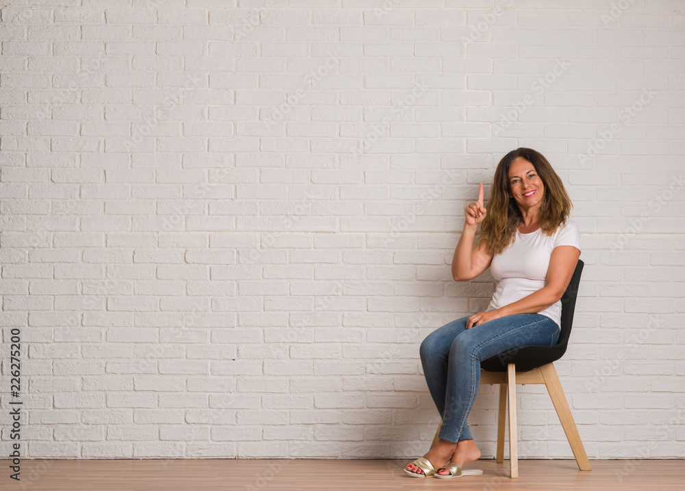 Middle age hispanic woman sitting on chair over white brick walll surprised with an idea or question pointing finger with happy face, number one Middle age hispanic woman sitting on chair over white brick walll surprised with an idea or question pointing finger with happy face, number one