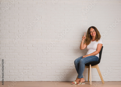 Middle age hispanic woman sitting on chair over white brick walll surprised with an idea or question pointing finger with happy face, number one