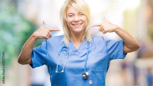 Young beautiful blonde doctor woman wearing medical uniform over isolated background smiling confident showing and pointing with fingers teeth and mouth. Health concept.