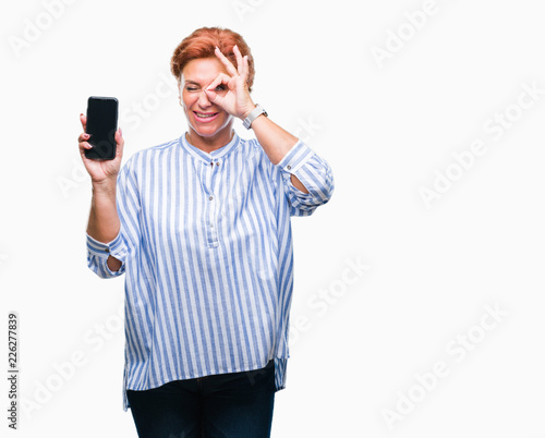 Atrractive senior caucasian redhead woman showing smartphone screen over isolated background with happy face smiling doing ok sign with hand on eye looking through fingers © Krakenimages.com