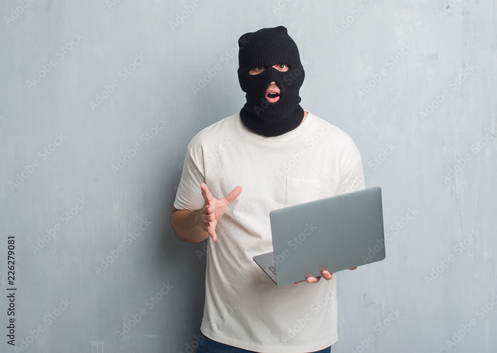 Young caucasian hacker man over grey grunge wall doing cyber attack using laptop scared in shock with a surprise face, afraid and excited with fear expression Young caucasian hacker man over grey grunge wall doing cyber attack using laptop scared in shock with a surprise face, afraid and excited with fear expression