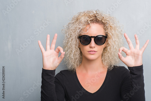Young blonde woman with curly hair over grunge grey background relax and smiling with eyes closed doing meditation gesture with fingers. Yoga concept.