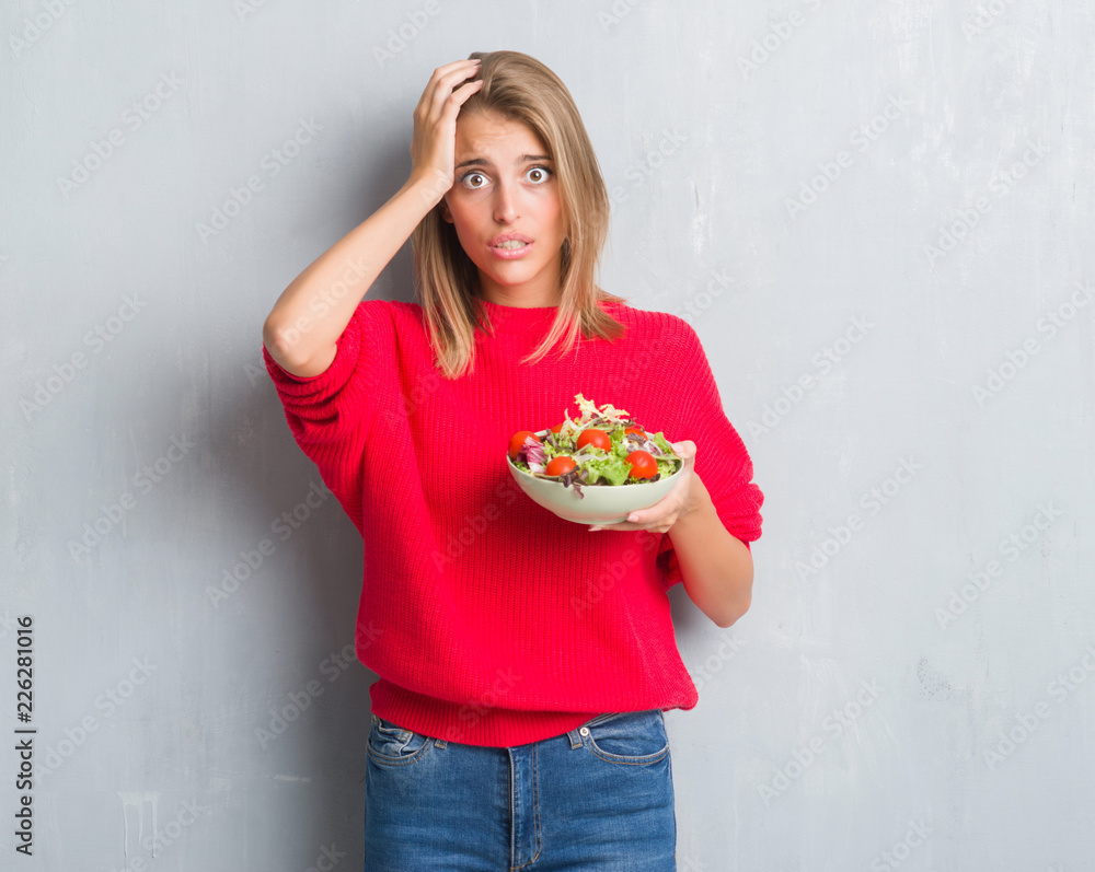 Beautiful young woman over grunge grey wall eating tomato salad stressed with hand on head, shocked with shame and surprise face, angry and frustrated. Fear and upset for mistake.