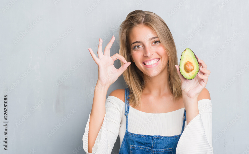 Beautiful young woman over grunge grey wall eating avocado doing ok sign with fingers, excellent symbol Beautiful young woman over grunge grey wall eating avocado doing ok sign with fingers, excellent symbol