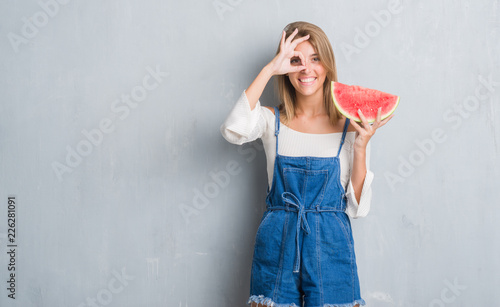 Beautiful young woman over grunge grey wall eating water melon with happy face smiling doing ok sign with hand on eye looking through fingers