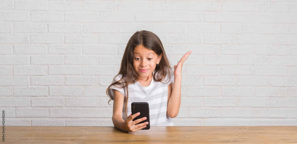Young hispanic kid sitting on the table using smartphone very happy and excited, winner expression celebrating victory screaming with big smile and raised hands Young hispanic kid sitting on the table using smartphone very happy and excited, winner expression celebrating victory screaming with big smile and raised hands