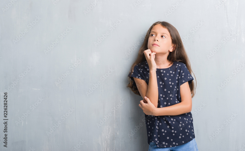 Fototapeta premium Young hispanic kid over grunge grey wall with hand on chin thinking about question, pensive expression. Smiling with thoughtful face. Doubt concept.