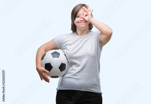 Young adult woman with down syndrome holding soccer football ball over isolated background with happy face smiling doing ok sign with hand on eye looking through fingers