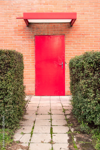 Vertical image. Red door surrounded by orange brick wall. An aile leads to the door and green busches are on both sides of the aile. Zlin, Czech Republic. photo