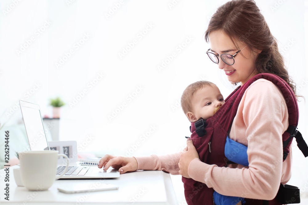 Young woman with baby in ergo carrier using laptop indoors Young woman with baby in ergo carrier using laptop indoors