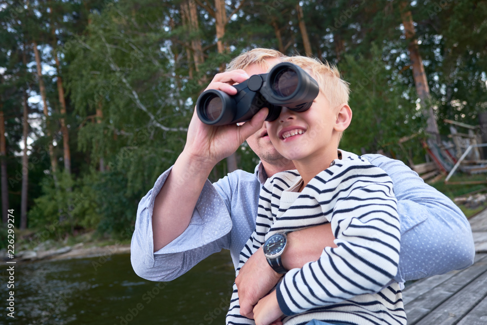 Boy in striped vest sitting with his father near the river or lake with binoculars in the background of the woods Boy in striped vest sitting with his father near the river or lake with binoculars in the background of the woods