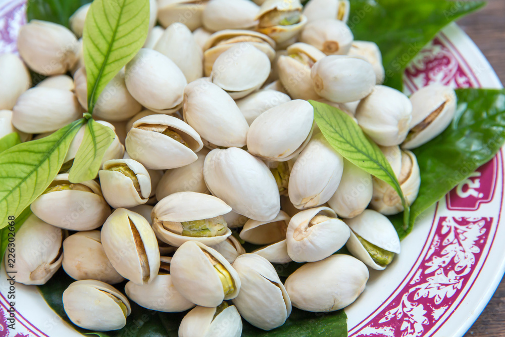 Close up of Pistachios nuts on wooden table. Pistachio in wooden bowl in background with green leaves Close up of Pistachios nuts on wooden table. Pistachio in wooden bowl in background with green leaves