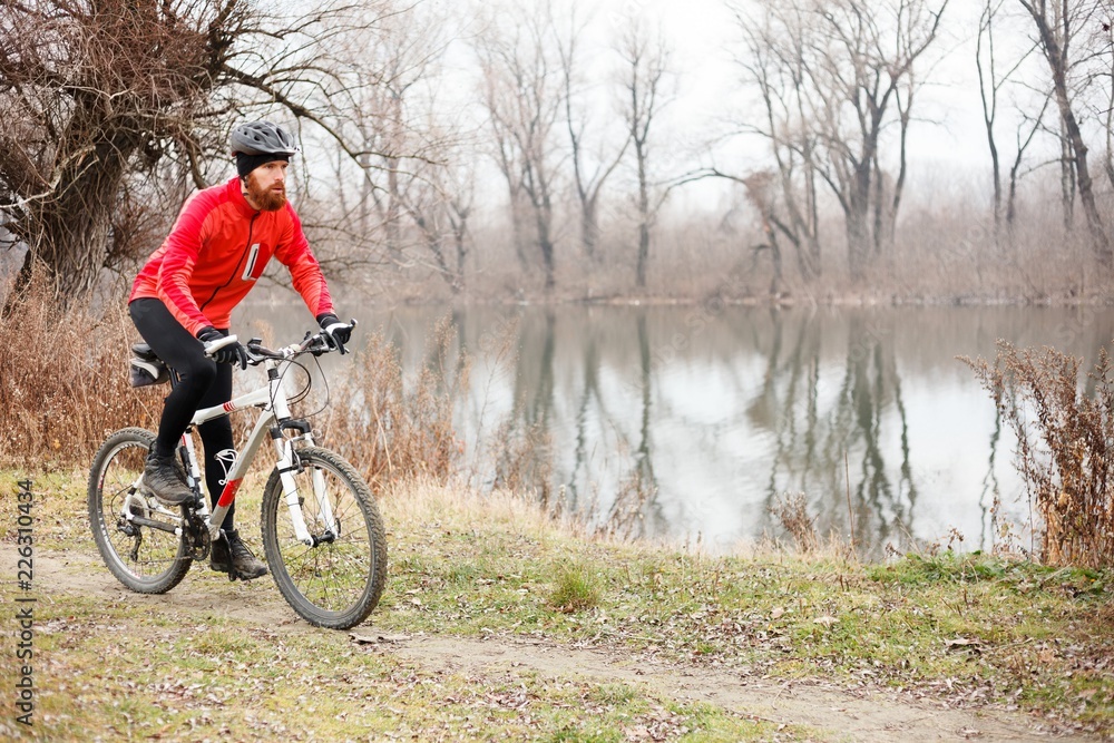 Determined young bearded man riding mountain bike by the river on a foggy autumn or winter day