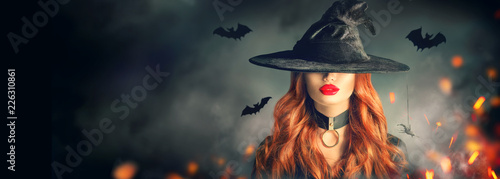 Halloween. Sexy witch portrait. Beautiful young woman in witches hat with long curly red hair over spooky dark magic forest background