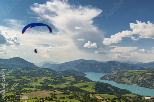 Bright paraglider tandem wing fly over beautiful mountain valley with green hills and blue lake.