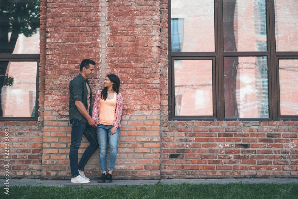 Urban style. Cheerful couple of young relaxed people standing next to the brick wall and smiling while holding hands and talking