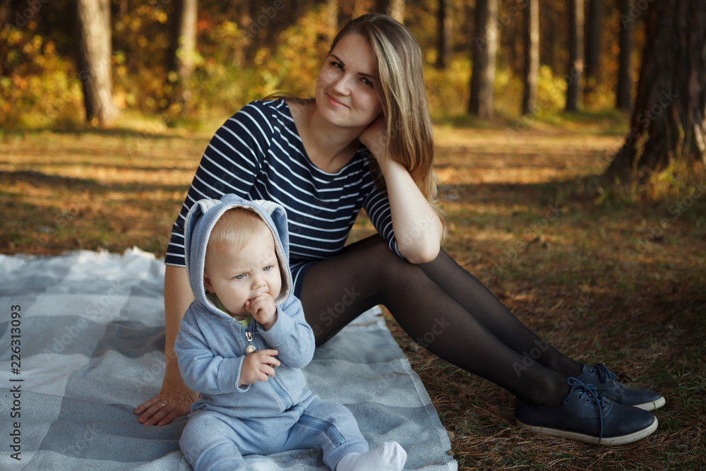 Woman with her son dressed in a blue rabbit suit are sitting on a plaid in the autumn forest. Woman with her son dressed in a blue rabbit suit are sitting on a plaid in the autumn forest.