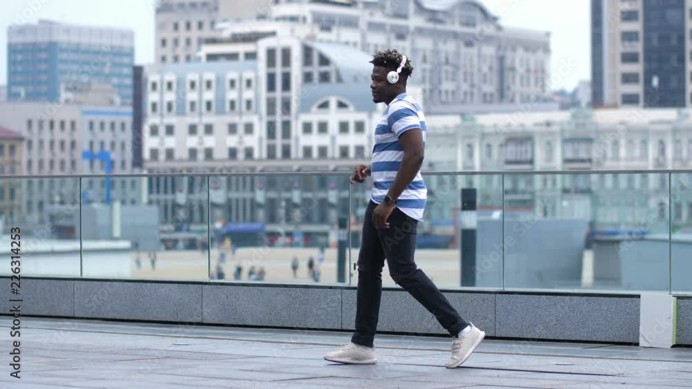 Cool trendy african american guy in headphones performing afrohouse dance on city street. Happy black male hipster dancing in afrobeat style outdoors over urbanscape background. Slow motion. Cool trendy african american guy in headphones performing afrohouse dance on city street. Happy black male hipster dancing in afrobeat style outdoors over urbanscape background. Slow motion.