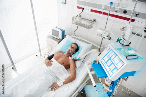 Top view portrait of middle aged gentleman on mechanical ventilator sleeping in recovery room after surgery. Man is lying with intravenous drip in his arm and pulse oximeter on finger photo