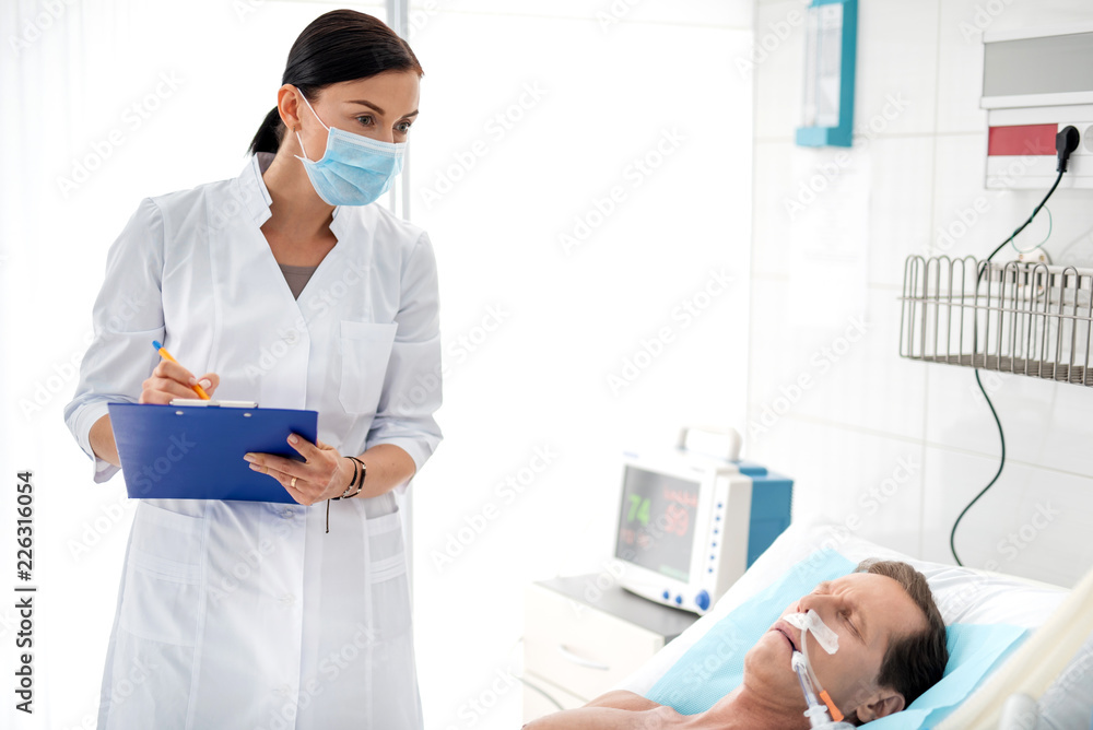 Waist up portrait of young lady in protective mask and white lab coat noting diagnosis of middle aged man. Sleeping gentleman lying in bed with endotracheal tube in mouth Waist up portrait of young lady in protective mask and white lab coat noting diagnosis of middle aged man. Sleeping gentleman lying in bed with endotracheal tube in mouth