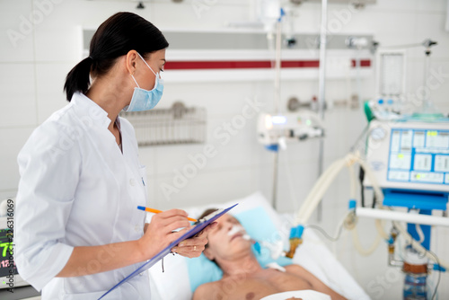 Waist up portrait of female medical worker monitoring condition of sick middle aged man. Gentleman lying in bed with endotracheal tube in mouth on blurred background