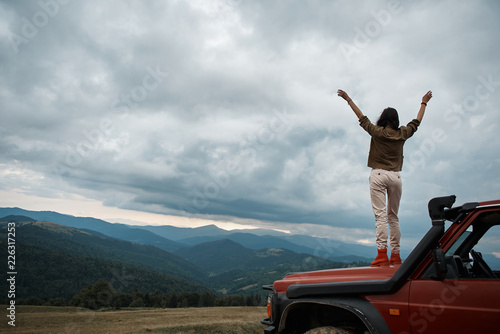 Feel free. Rear view of a young slim female tourist standing in the car hood while enjoying view over the mountains