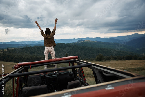 Panoramic view of mountains covered with coniferous forest with a nice woman standing on the car hood