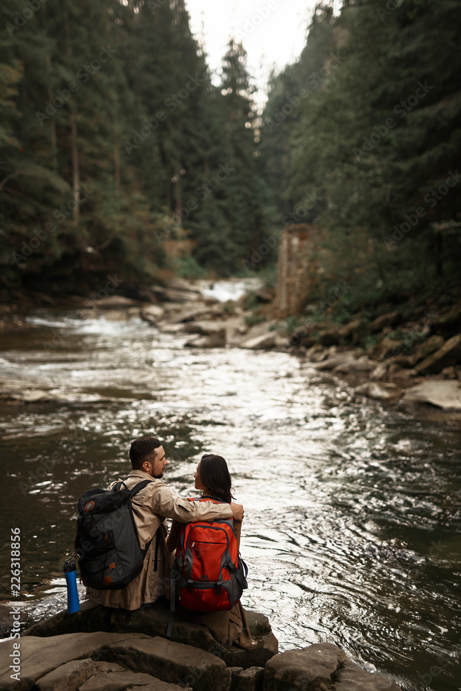 Peaceful stream. Kind loving man gently hugging his beloved woman while sitting near the river with her Peaceful stream. Kind loving man gently hugging his beloved woman while sitting near the river with her