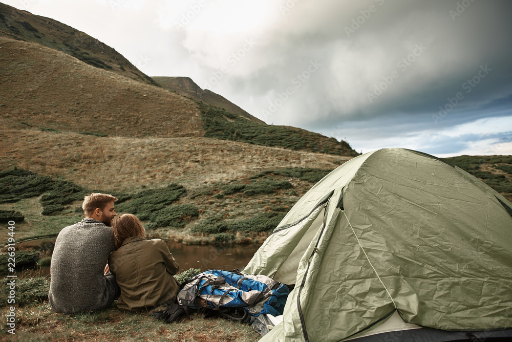Fototapeta premium Cuddling near tent. Peaceful young couple sitting near the tent and looking relaxed while enjoying beautiful landscape