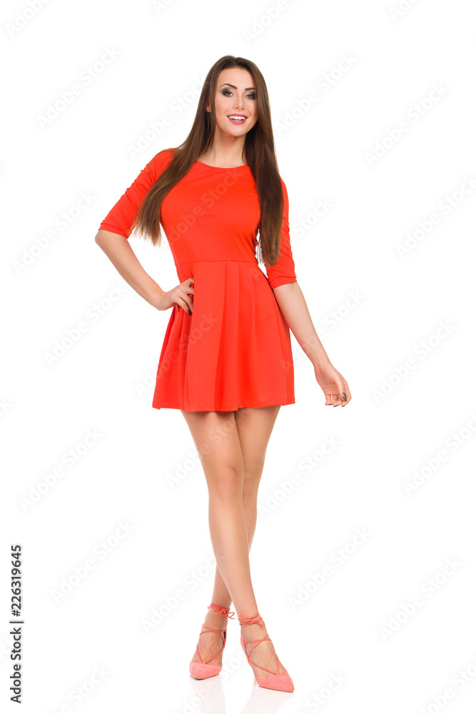 Smiling Fashion Model In Red Mini Dress And High Heels Is Standing With Legs Crossed Smiling Fashion Model In Red Mini Dress And High Heels Is Standing With Legs Crossed