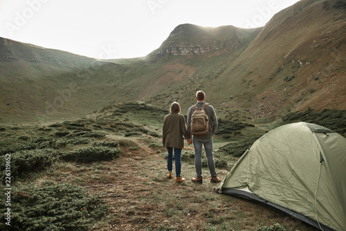 Couple near tent. Peaceful relaxed young man and woman standing near the tent and holding hands while looking at the landscape