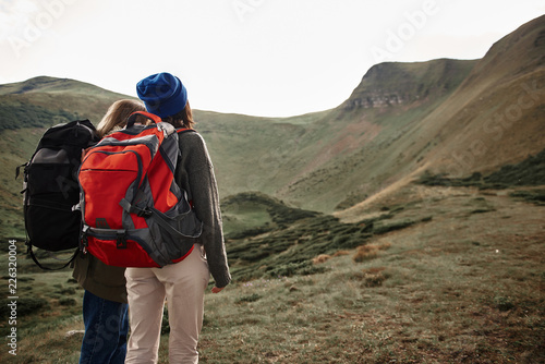 Admiring beauty. Two young women carrying big heavy backpacks and wearing comfortable clothes while traveling and looking at the landscape