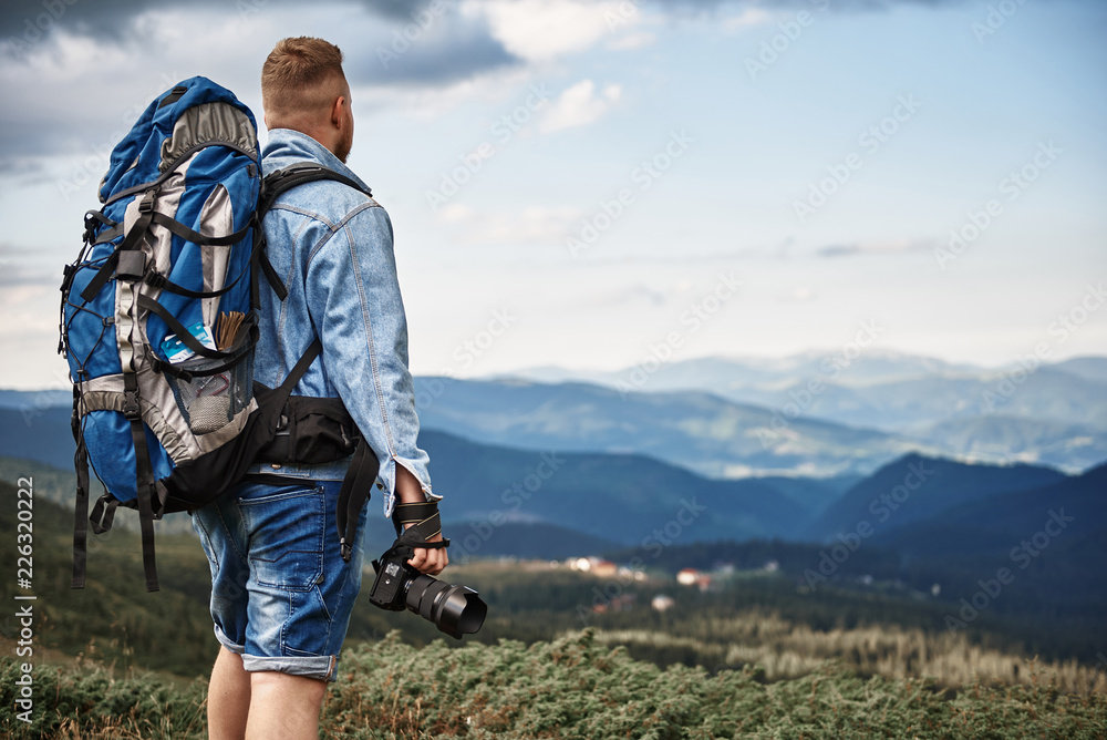 Photographer with backpack. Calm tall young photographer standing with modern camera and looking at the view while traveling in the mountains