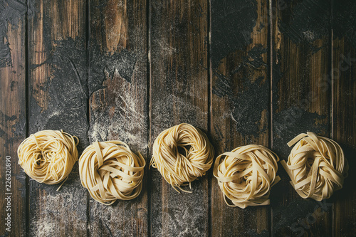 Variety of italian homemade raw uncooked pasta spaghetti and tagliatelle in row with semolina flour over dark plank texture wooden table. Flat lay, copy space