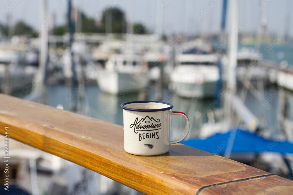 An aluminum enamel mug for a hike on a wooden table on the board of the yacht in the port of Italy among other yachts on a sunny summer day.