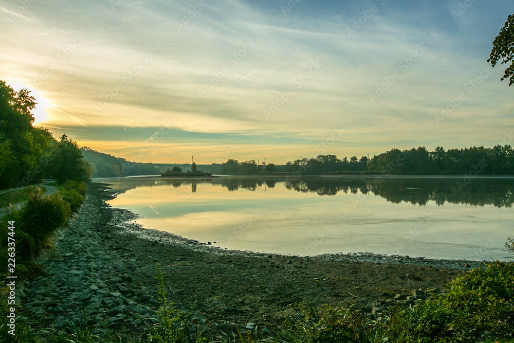 Fototapeta premium Morning view of sun dawn over a lake, reflection in water of blue, pink and yellow sky, dark lake shore in foreground, autumn landscape