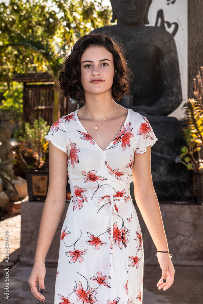Fototapeta premium Headshot of beautiful young teen dressed in long flower print dress walking along concrete pathl in garden.