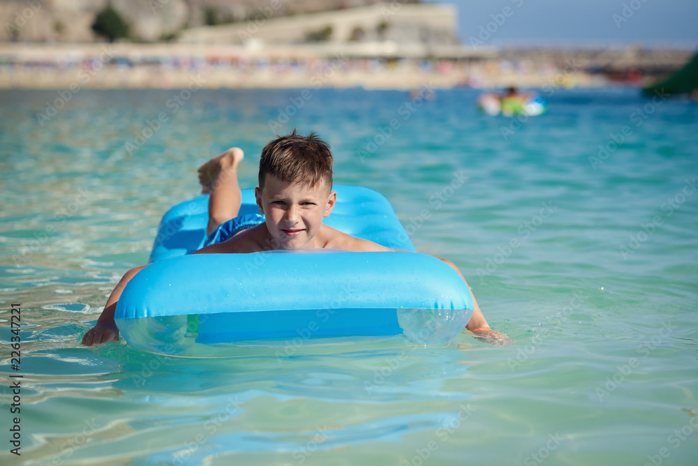 Happy European boy in striped swimming shorts is laying on a blue floater in the ocean. He is enjoying his holidays and smiling.