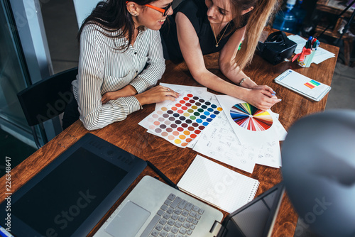 Two female architects working together using color swatches sitting at desk with laptop, graphic tablet in design studio