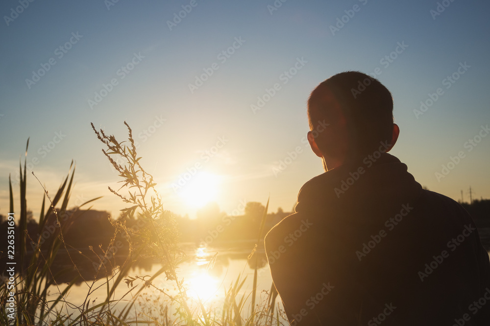 One Young man sitting on the grass by the lake at sunset. Evening autumn sunset.