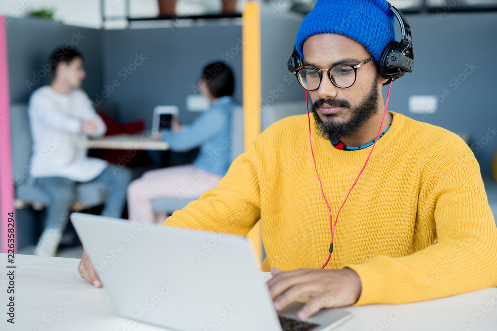 Serious concentrated young middle-eastern man with beard and mustache wearing headphones sitting at table and working with laptop Serious concentrated young middle-eastern man with beard and mustache wearing headphones sitting at table and working with laptop