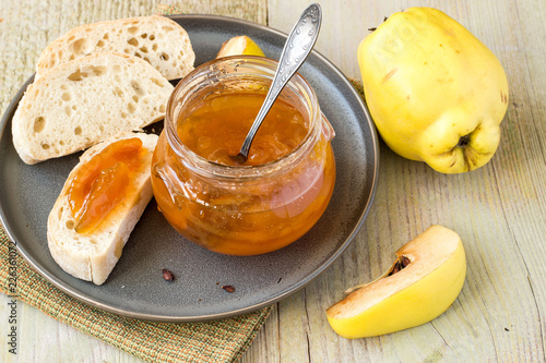 A small glass jar of jam, a fresh ripe quince fruits and homemade white bread on an old table. 