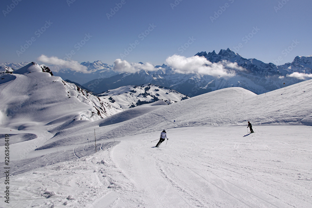 Skiers in French Alps carve turns as they make their way down a piste in Avoriaz, Portes du Soleil ski area. Swiss Alps can be seen in background