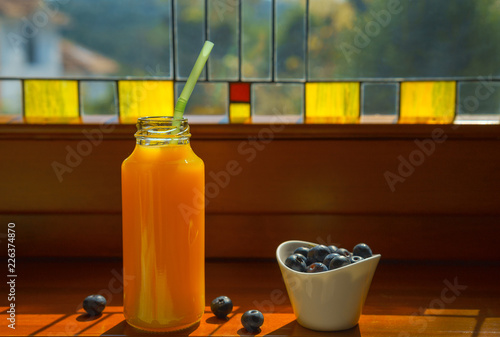 Still life with healthy breakfast ingredients orange juice in glass bottle and white bowl with blueberries stand by the colorful stained glass window. Organic. Wooden rustic background.