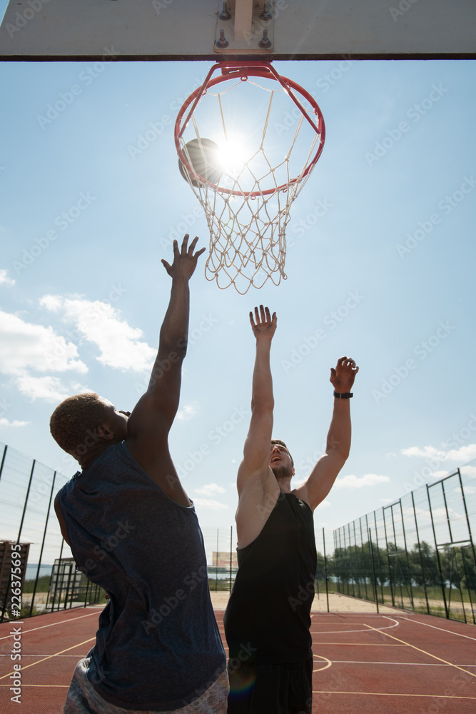 Naklejka premium Action portrait of two young men playing basketball and jumping by hoop against blue sky lit by sunlight