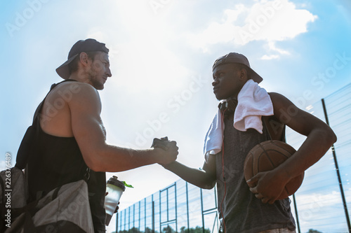 Side view portrait of two basketball players shaking hands against blue sky after match in outdoor court, copy space