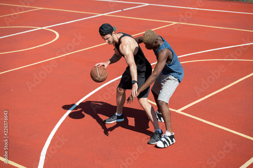 High angle action shot of two handsome muscular men, one of them African, playing basketball in outdoor court lit by sunlight, copy space