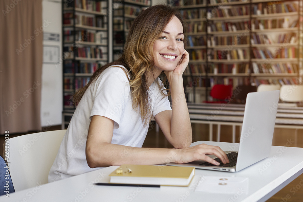 Young woman long hair analyst working at co-working office on laptop, bookshelf behind. Businesswoman learning alone in library. Young woman long hair analyst working at co-working office on laptop, bookshelf behind. Businesswoman learning alone in library.