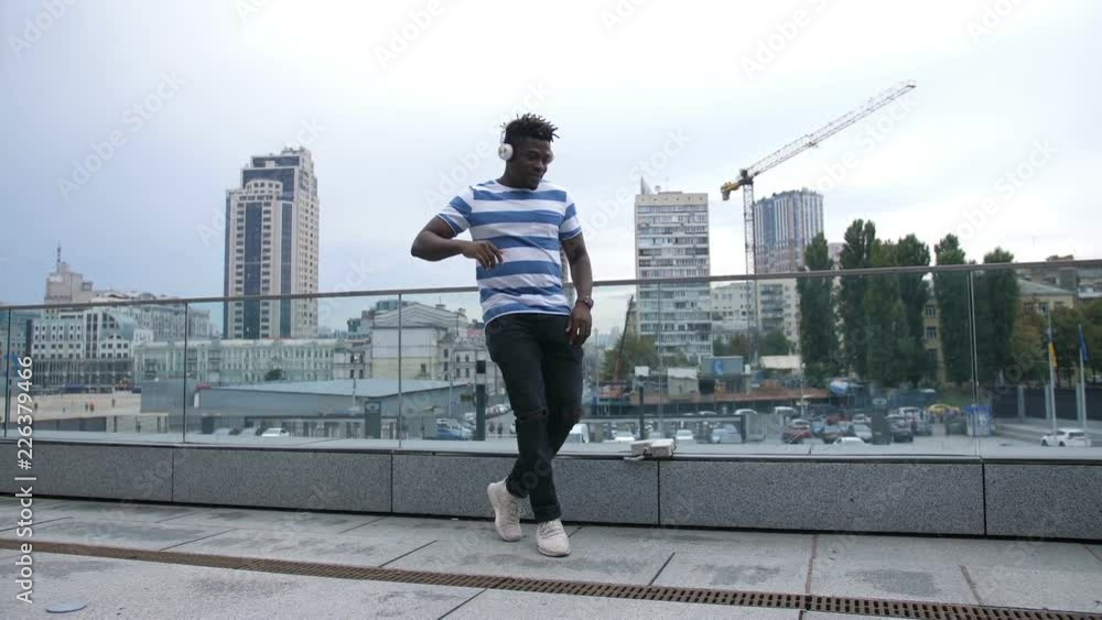 Handsome trendy african american hipster in white headphones walking along street and moving in afrohouse style dance over cityscape background. Cheerful black man dancing outdoors. Slo mo. Handsome trendy african american hipster in white headphones walking along street and moving in afrohouse style dance over cityscape background. Cheerful black man dancing outdoors. Slo mo.